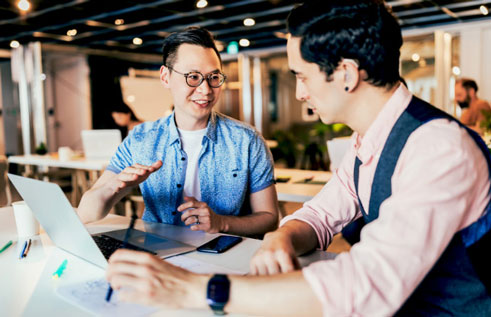 Two men sit at a desk and have a conversation