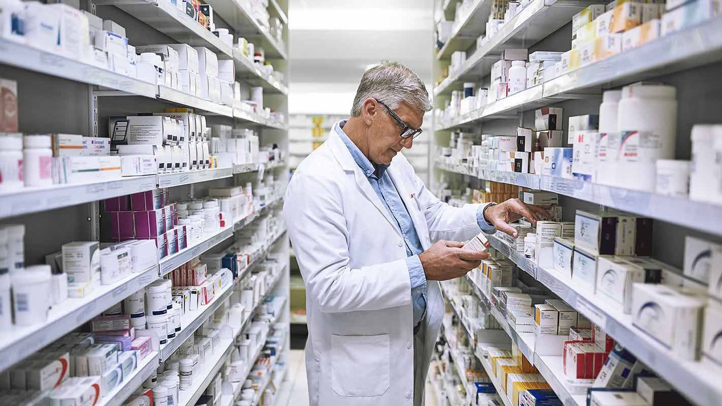 A pharmacist stands in the aisles, reading a medicine box