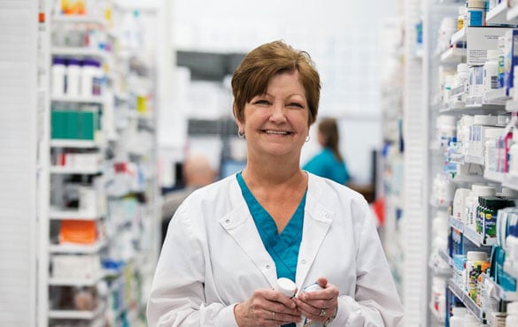 A female pharmacist smiles behind the counter, surrounded by medications on shelves