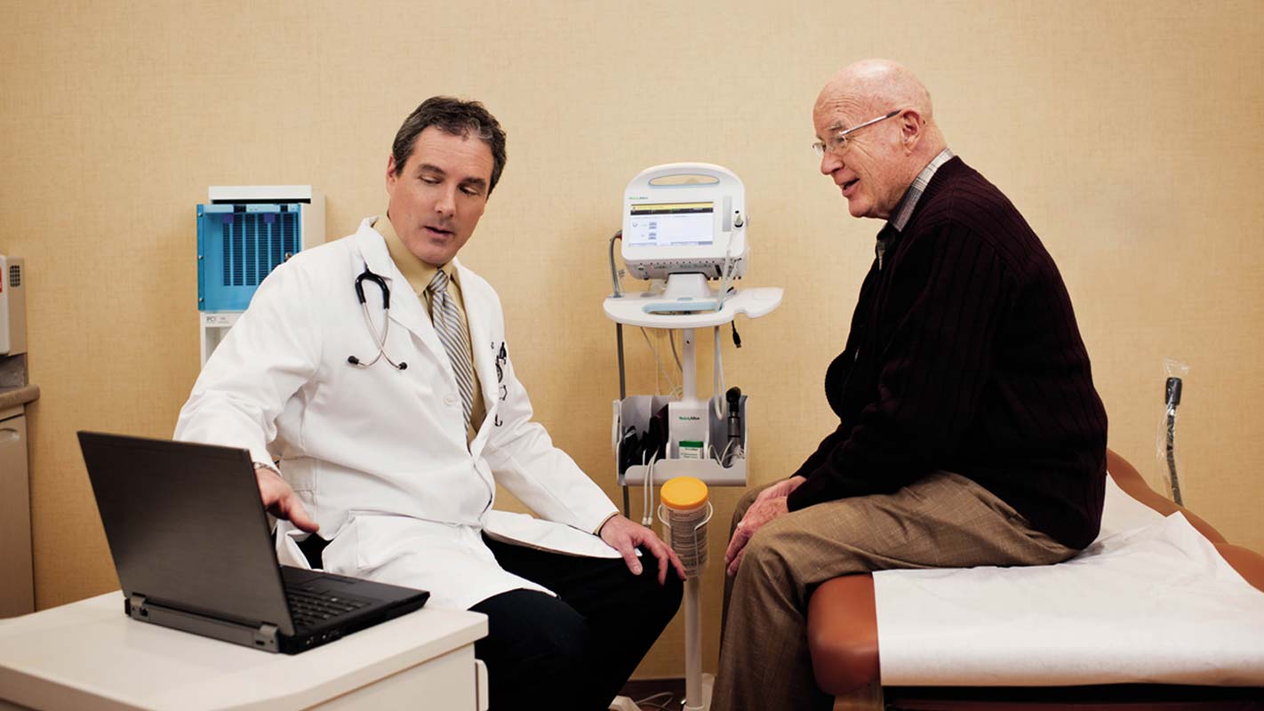 A doctor looks at a laptop as a patient sits on an exam table and listens