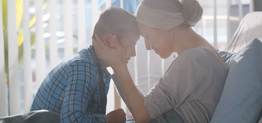 A female cancer patient sits in bed with a scarf around her head and embraces a young boy sitting across from her