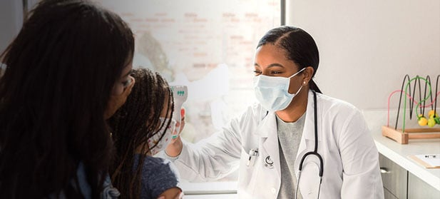 A physician uses a forehead thermometer to take a child's temperature while the child sits on her mother's lap