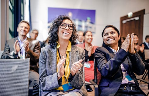 People sit together in folding chairs, clapping hands and smiling during a presentation