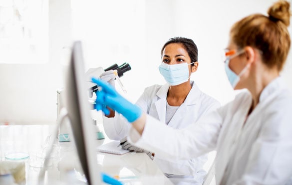 Two scientists sit at a table, one is using a microscope, the other a computer